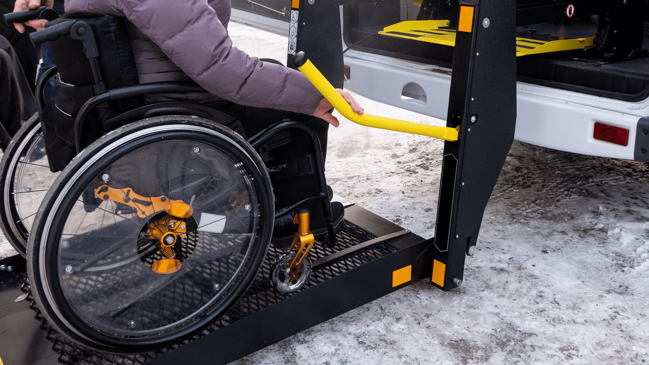 An accessible taxi van with a wheelchair lift is parked on a snowy residential street in winter. A man appears to be operating the mechanical wheelchair lift, which is lowered to pavement level. The lift is black metal and has a bright yellow handrail. On the lift is a woman in a wheelchair, bundled up in a coat. She waits as the man assists her into the specialized disability transportation vehicle.