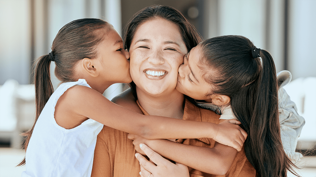 Portrait of an Asian mother and 2 daughters on each side of her to embrace her, with their arms around her. The mother is smiling with her teeth.