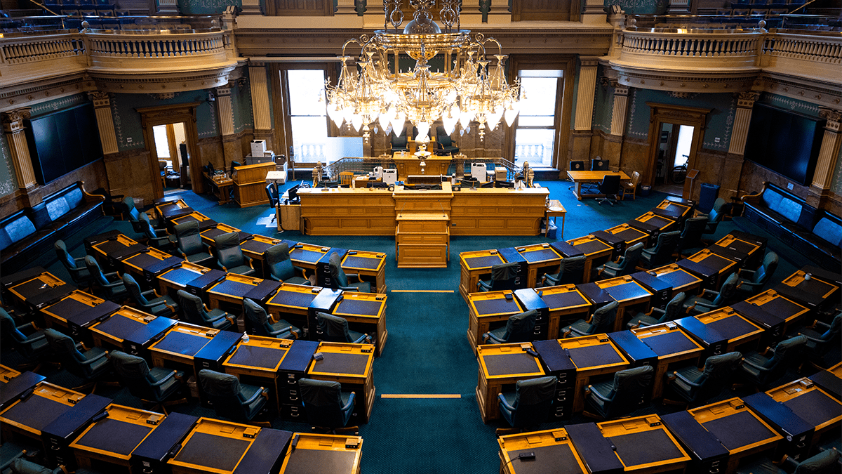 Inside the Colorado Capitol in the House or Senate floor with several desks arced and facing the main podium.