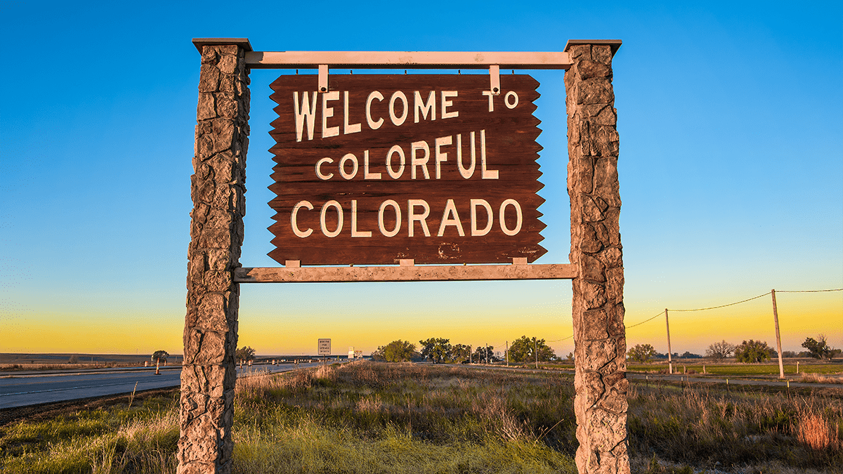 Welcome to colorful Colorado wooden street sign. The sign is held together by two rock pillars. Blue and yellow gradient sky in the background.