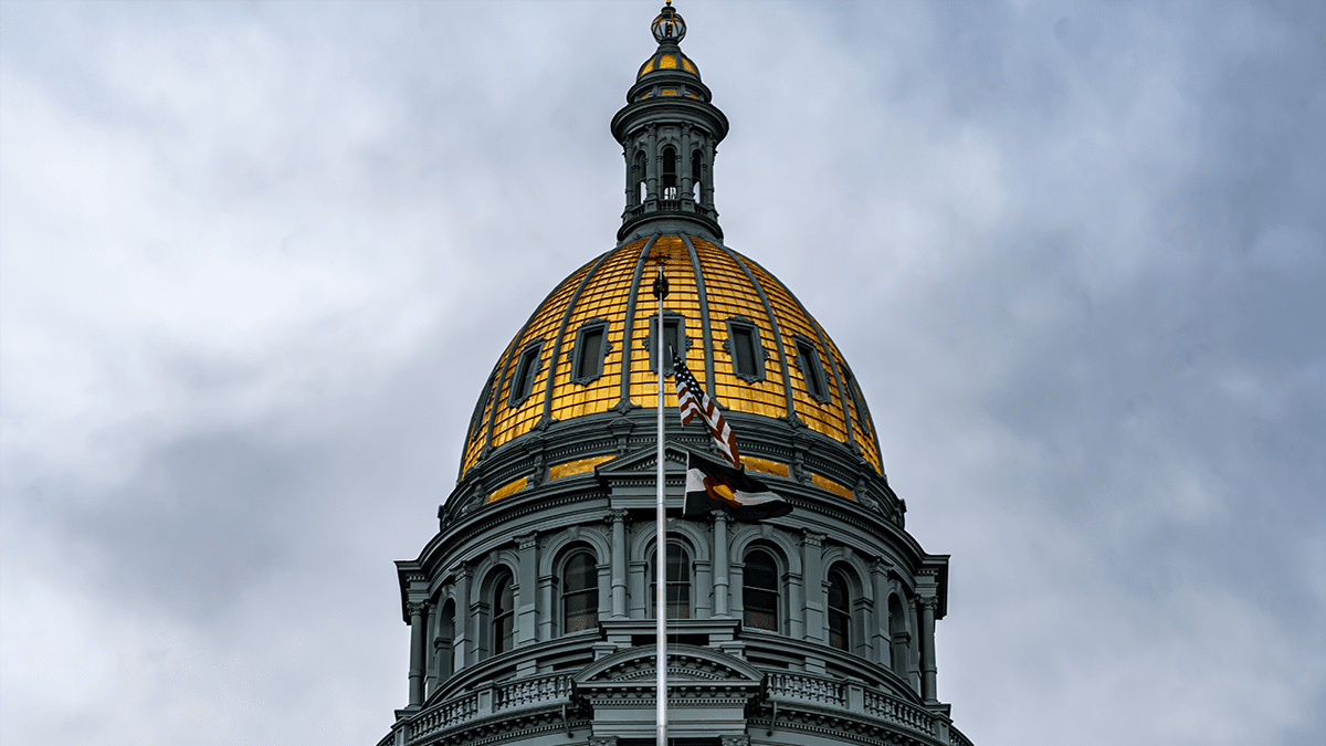 The gold top of the Colorado Capitol building with the American and Colorado state flags flying. The sky is cloudy and has an ominous tone to the photo.
