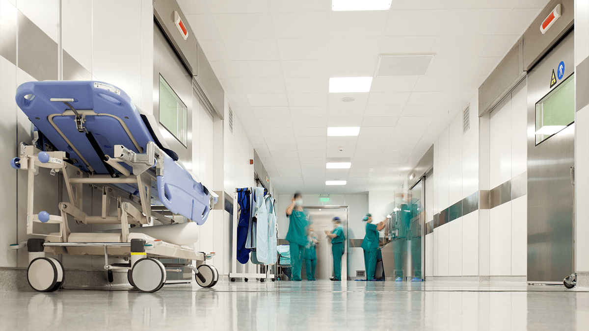 Hospital surgery corridor. Gurney on the left of the image with no one on it. Doctors are in the background in teal scrubs.