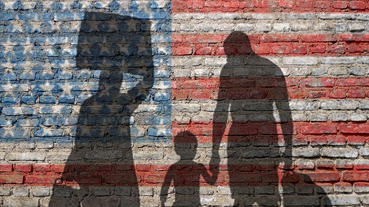 A brick background painted as the American flag with shadows of an immigrant family with a child and carrying their belongings.