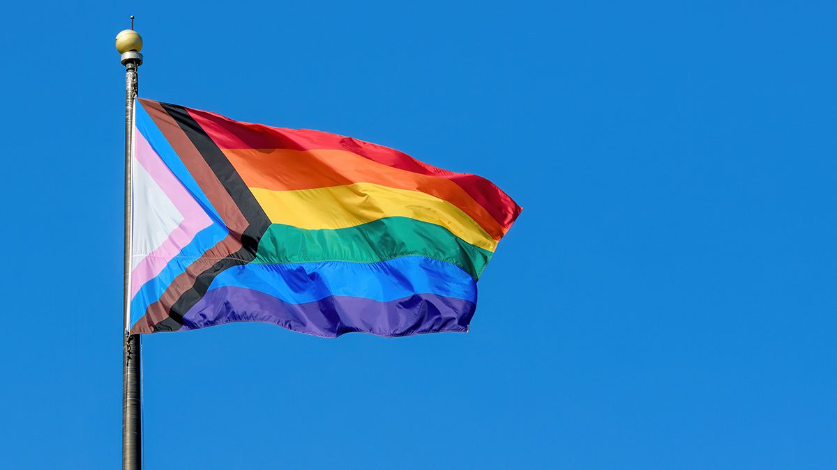 A LGBTQ+ rainbow progress pride flag flaps from a flagpole in a brisk breeze. Blue sky behind.