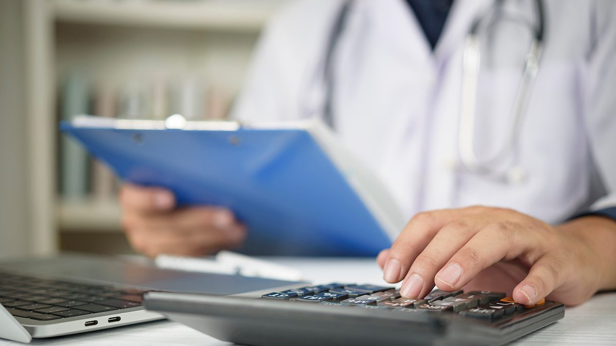 Close up of doctor hand using a calculator and holding a clipboard with medical paperwork. Concept of healthcare costs, medical insurance premiums, hospital fees, and financial administration.