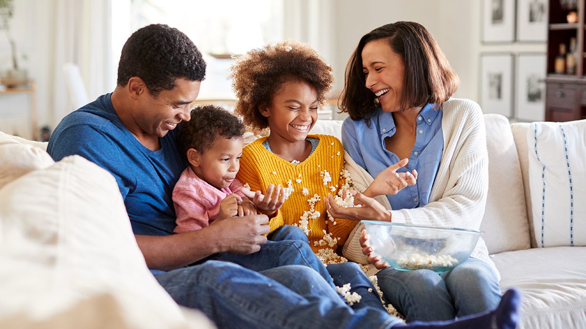 Young mixed race family sitting together on the sofa in their living room watching a scary movie, clearing up spilled popcorn, selective focus