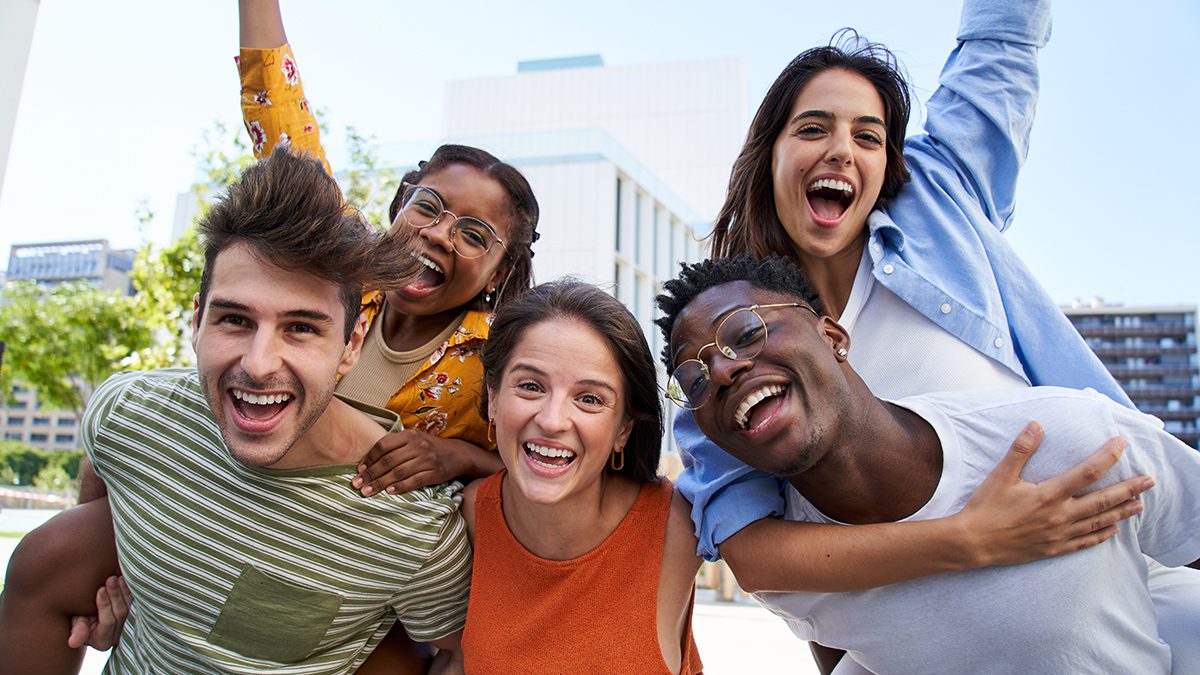 Portrait of happy young friends looking at camera excited. Smiling group of classmates having fun together outdoors. Crazy community of college students. Multicultural people hugging and piggyback.