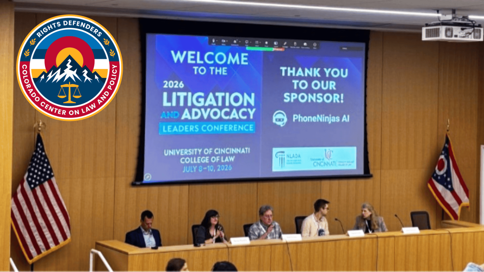 Litigation conference leaders sit at a panel in front of a room with a screen behind them. Flags on either side of them with CCLP Rights Defenders logo in the top left corner.